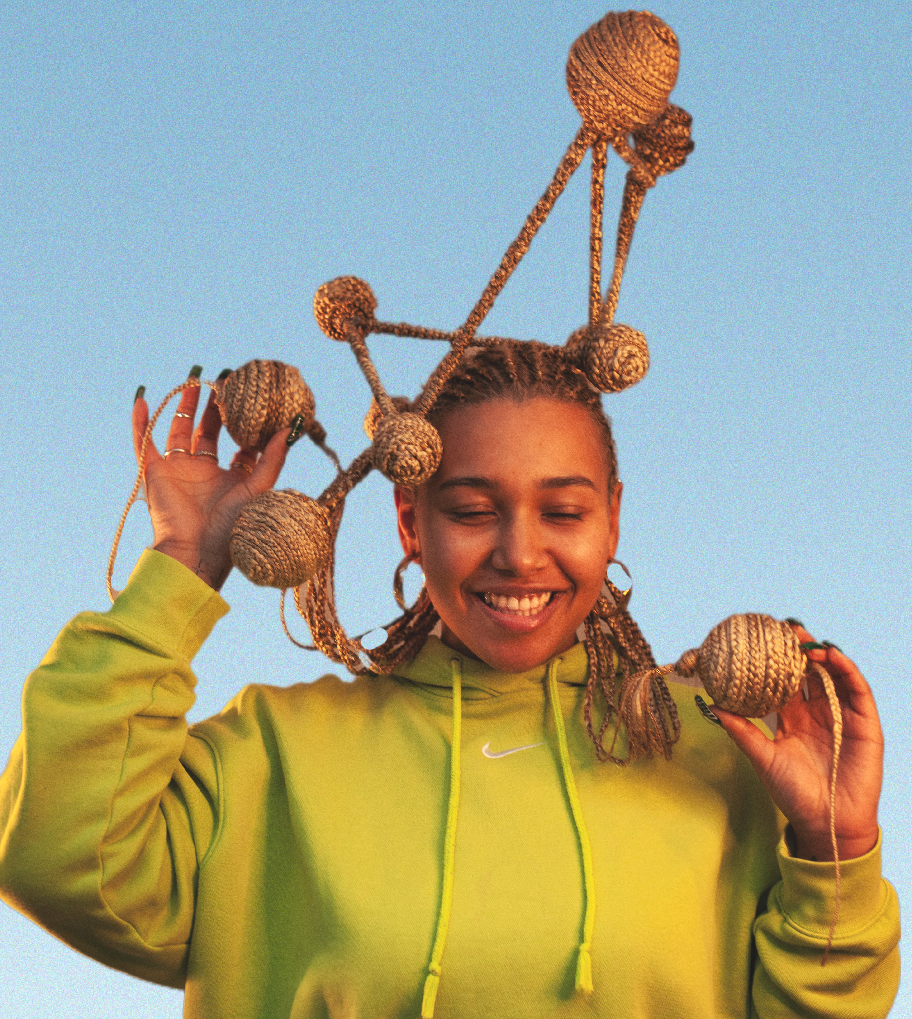 A smiling young brown woman with her eyes shut, wearing a bright green tracksuit with a brown/golden cornrow hairstyle surmounted by a braided brown/gold coloured structure composed of braided interconnected lines having a braided sphere at the nodes of the linear braids. The model also has a brown/gold braided sphere each in her hands. 