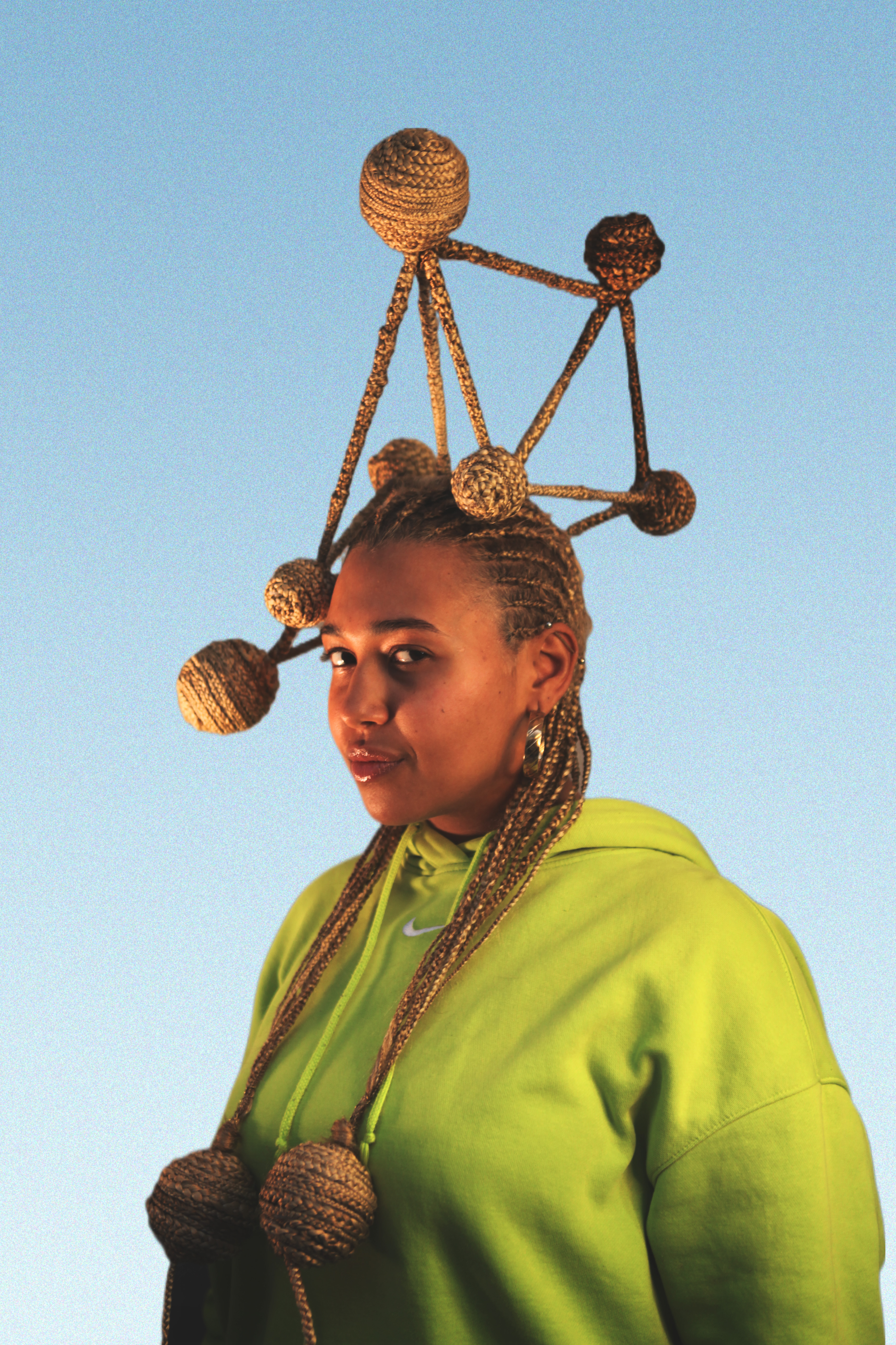 A young brown woman wearing a bright green tracksuit. She is smiling and standing with her side slightly turned to the camera. She has a golden/brown cornrow hairstyle. On her head is a golden/brown structure composed of braided lines and braided spheres at the node of the lines. Some of the braided spheres are attached to the model's cornrows and fall over her shoulders.