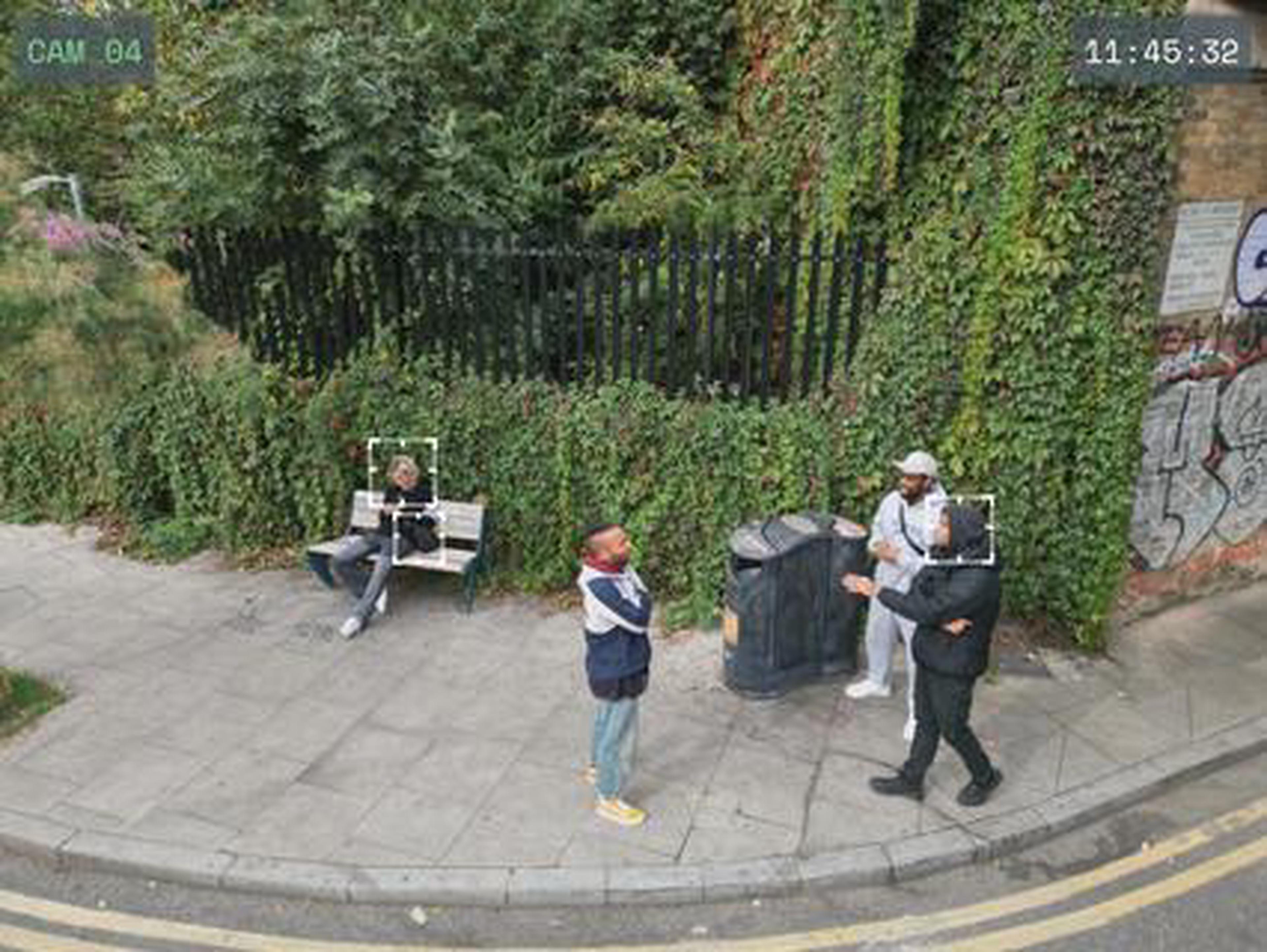 Landscape shaped image. Four people are outside on a pavement in front of some railings and lush green foliage. Two of them have a white frame superimposed round their heads, showing them being surveilled, perhaps from a drone. 