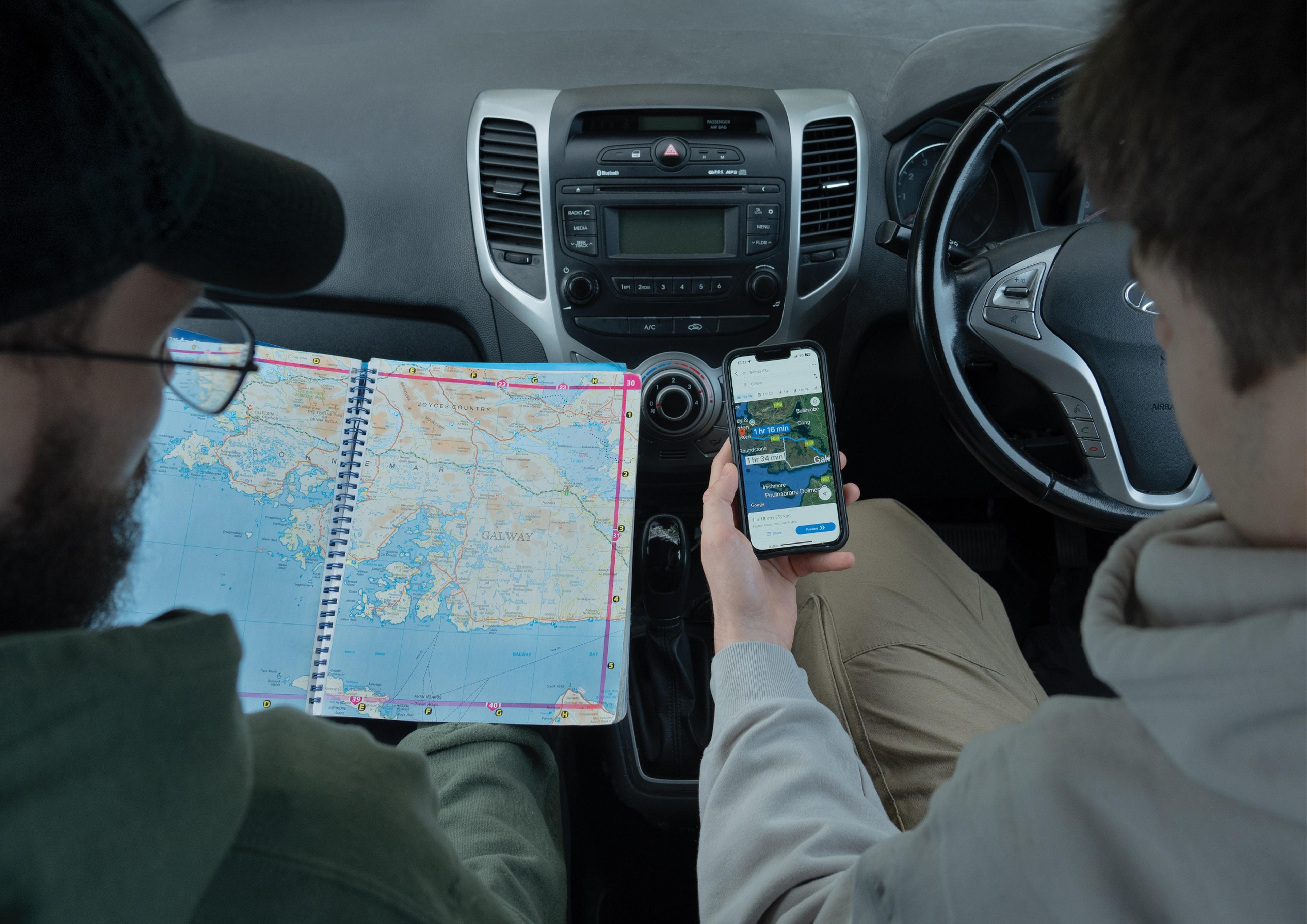 Two men are seated in the front seats of a car and the image is from the perspective as looking over the shoulders of the two individuals. The driver, on the right, holds a smartphone showing Google Maps with a route from Galway to Clifden, displaying the travel time and directions. The passenger, on the left, wears glasses and a dark green hoodie and holds a large, spiral-bound road atlas open to a page showing the west coast of Ireland. The car’s dashboard and controls are visible between them. 